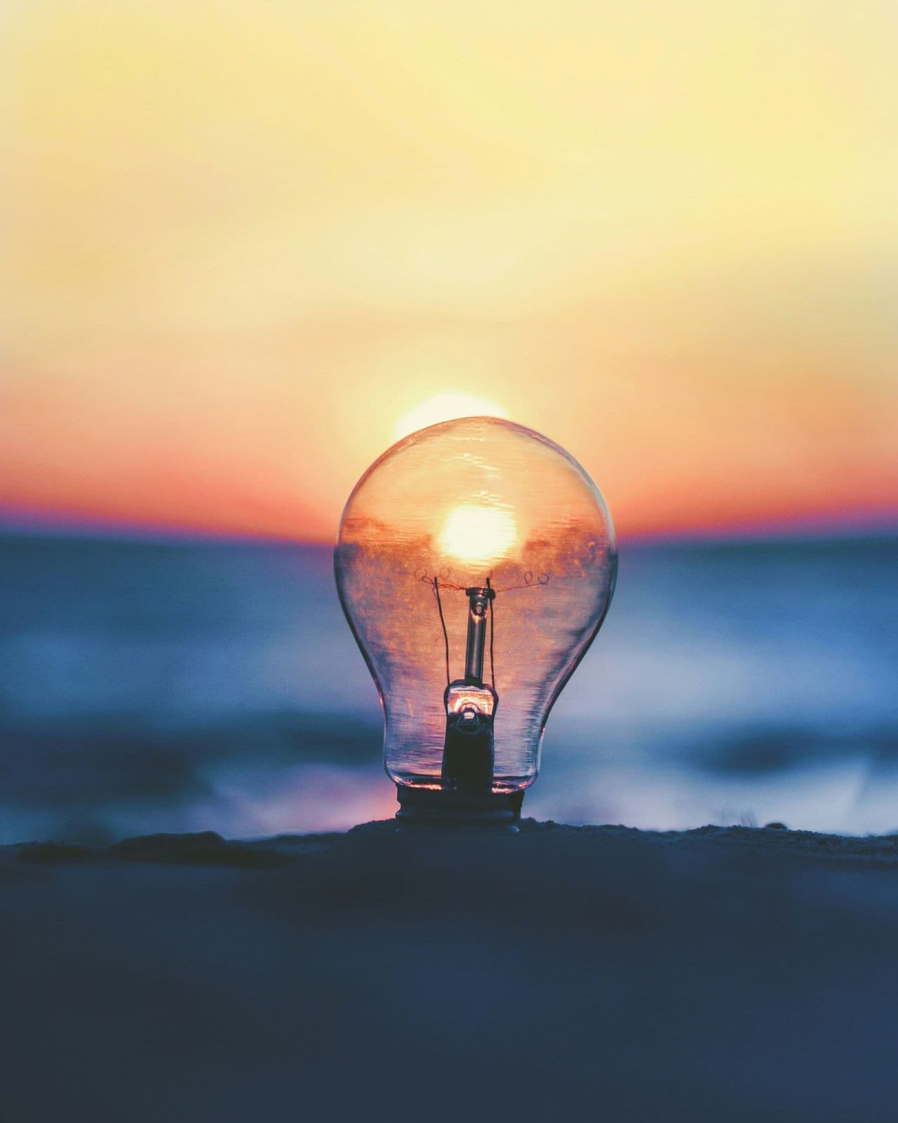 Lightbulb resting on the shore at sunset, glowing against the sea