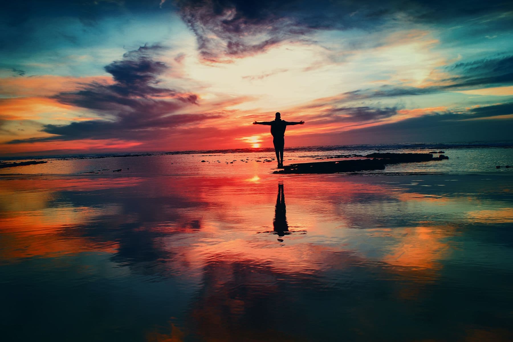 Man standing with arms open at sunset, reflected in still water